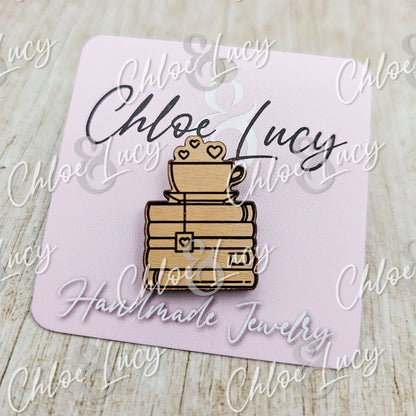 Wooden pin featuring a teacup atop stacked books with hearts, displayed on a pink card.