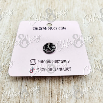 Small silver peace sign enamel pin on pink card, minimalist style, wood background